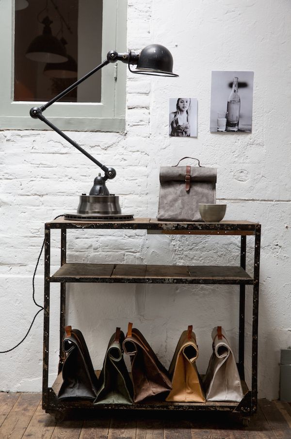 A stylish industrial setup with a black metal desk lamp and a small collection of items, including a gray bag and a bowl, on a distressed metal shelving unit. The lower shelf holds neatly rolled-up canvas bags in various colors, creating a functional and visually appealing display.