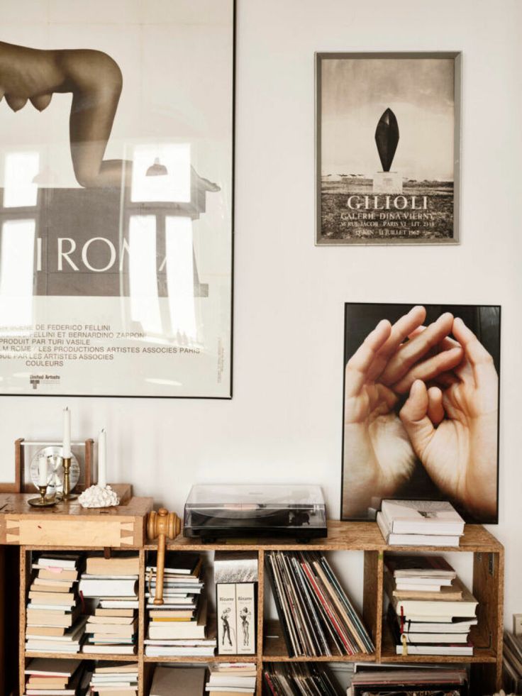An eclectic living area with a vintage green metal cart used as a side table. The cart is filled with books and topped with decorative items, including a nativity scene and a white candle. The wall behind features white paneling and large windows with transom lights, allowing natural light to flood the space. Two small art pieces, including a sketch and a photograph, are hung on the wall.