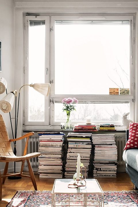 A cozy living room with large windows that allow natural light to flood the space. The room includes a stack of magazines serving as a side table, a wooden chair, and a modern floor lamp, creating a warm and inviting atmosphere.