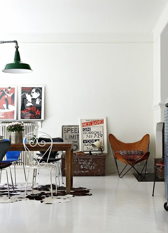 A dining area with eclectic decor, featuring a vintage-style metal dining set, a butterfly chair with a leather seat, and various framed posters and signs leaning against the wall. The room has a bright and airy feel with white walls and a mix of modern and retro elements.
