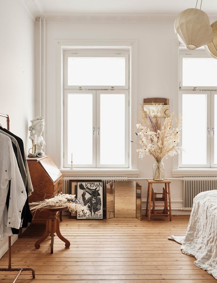 A bright room with large windows letting in abundant natural light. The room features a wooden floor, a small wooden table with dried flowers, a vintage desk, and minimalist decor elements.