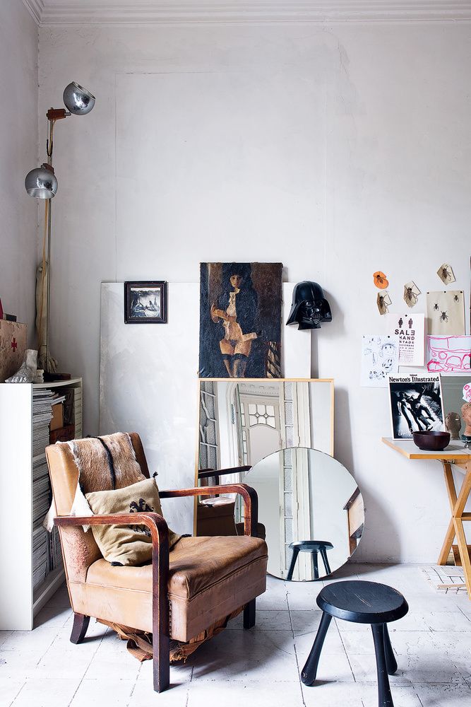 An eclectic living area with a mix of vintage and contemporary decor. A large portrait and various framed artworks are casually leaned against the wall. The room includes a vintage armchair with a plush cushion, a small black stool, a round mirror, and unique lighting fixtures.
