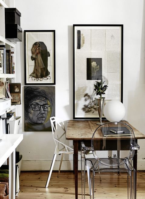A cozy corner of a room with a wooden table and a clear acrylic chair. The wall is decorated with three edgy art pieces: a portrait of a person’s face, an abstract composition, and a framed artwork with a mixture of textures and images. A small vase with flowers and a white spherical lamp sit on the table, adding a touch of elegance to the space. Shelves filled with books and storage boxes are visible to the left.