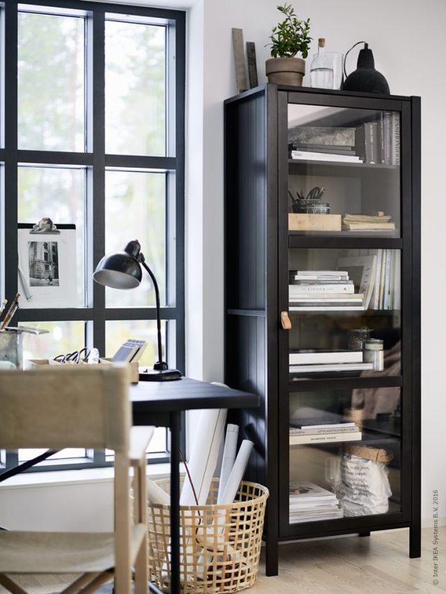 A modern, minimalistic home office with a black metal and glass cabinet filled with neatly arranged books and decor items. A wooden desk with a black lamp and a basket holding rolled-up papers sits next to a large window, providing ample natural light and a clean, organized workspace.