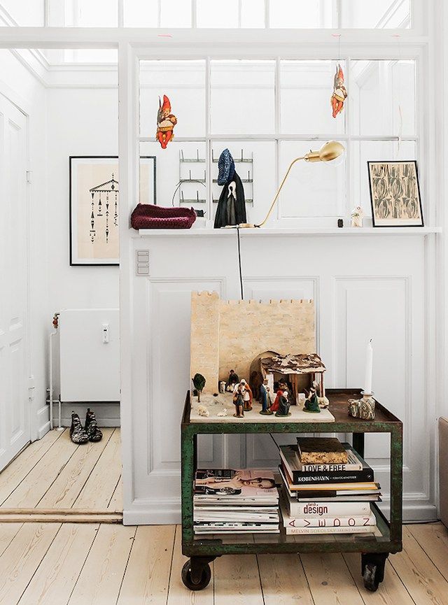 A stylish kitchen nook with a vintage green rolling cart used as a side table, displaying various books and decorative items. The white walls, large windows, and wooden floors create a bright and inviting atmosphere, complemented by unique wall art and accessories.