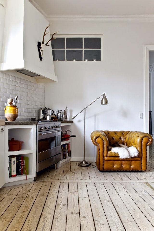 A cozy kitchen corner featuring a mustard yellow leather armchair with a plush throw, placed next to a stainless steel stove and white subway tile backsplash. The rustic wooden floor and tall floor lamp add warmth and charm to the space.