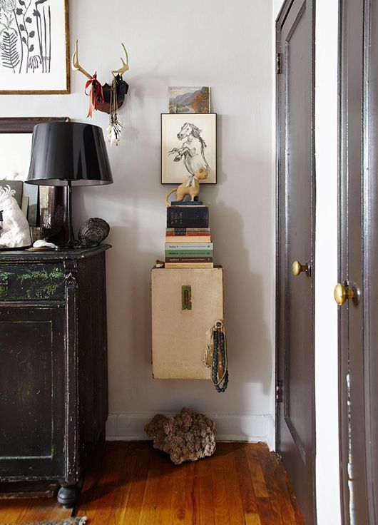 A stylish hallway corner featuring a stack of books on a vintage suitcase, topped with a small animal figurine. The vignette includes a black table lamp, framed art, and decorative antlers on the wall, creating an eclectic and artistic look.