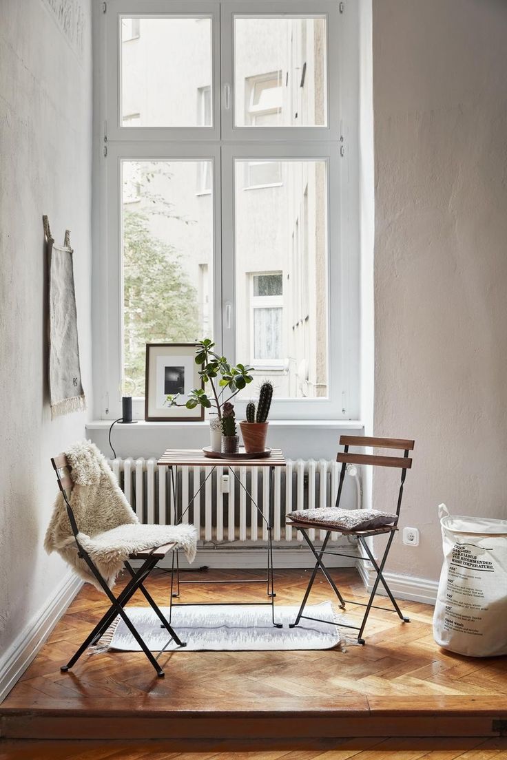A small, bright corner with a large window bringing in natural light. The space is furnished with a small table and two chairs, potted plants, and minimalist decor, creating a serene and airy nook.