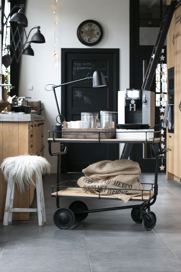 Industrial style coffee station in a home office, featuring a metal and wood cart with a coffee machine, various metal containers, and a burlap sack. The area is accented with a black desk lamp, a white furry stool, and a black door in the background.