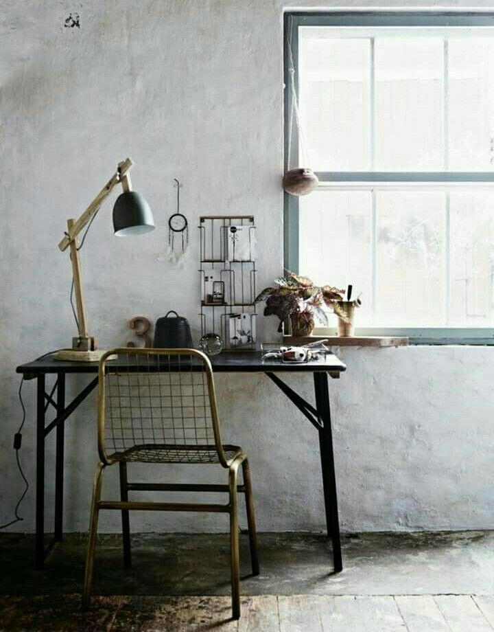 A minimalist boho home office with a black metal desk and a wire metal chair. The desk is placed next to a large window that brings in natural light, adorned with a wooden desk lamp and small decor items. The setting features a rustic, textured wall and simple, organic decor.