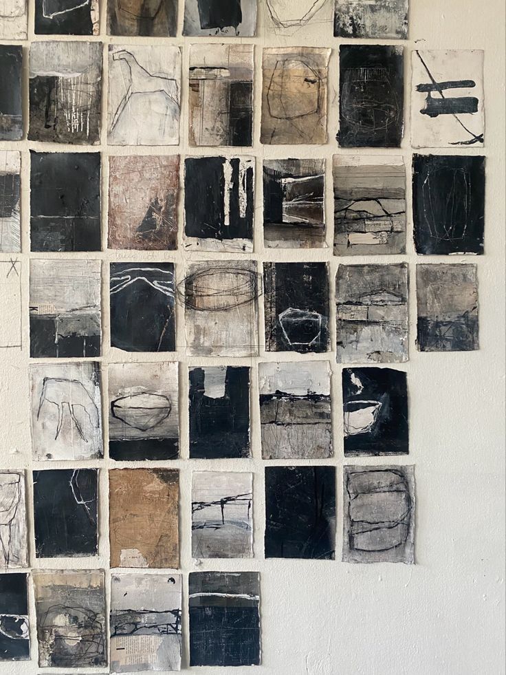 A wall decorated with a grid of small abstract artworks in various shades of black, white, and brown. Each piece features unique designs and textures, contributing to a cohesive yet diverse visual display in an art studio setting.