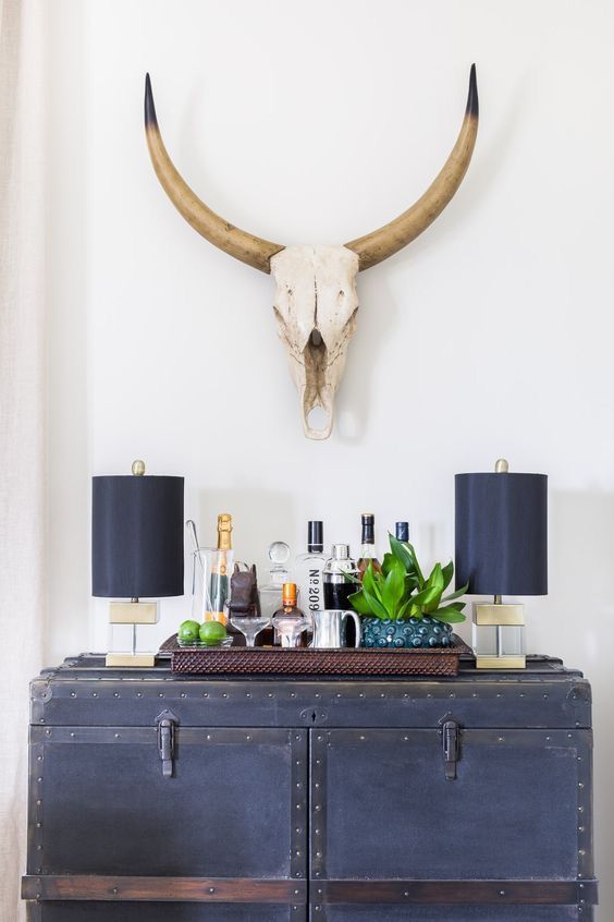 A stylish home bar setup featuring a vintage black trunk with two modern black table lamps, a decorative tray with bottles and glasses, and a mounted skull with large horns on the white wall.