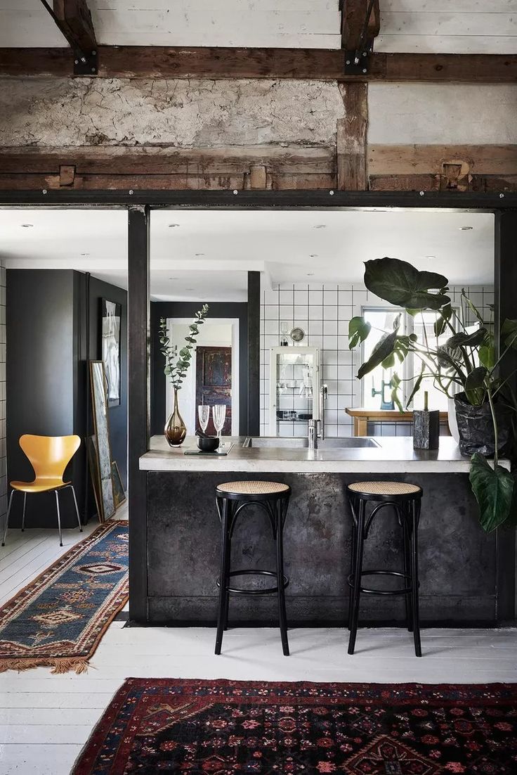 A boho-style kitchen featuring a mix of rustic and modern elements. The space includes a large island with a concrete countertop, two black stools with rattan seats, exposed wooden beams, and a textured, weathered wall. The kitchen is accented with a large potted plant, decorative vases, and a patterned rug, creating a warm, eclectic atmosphere.