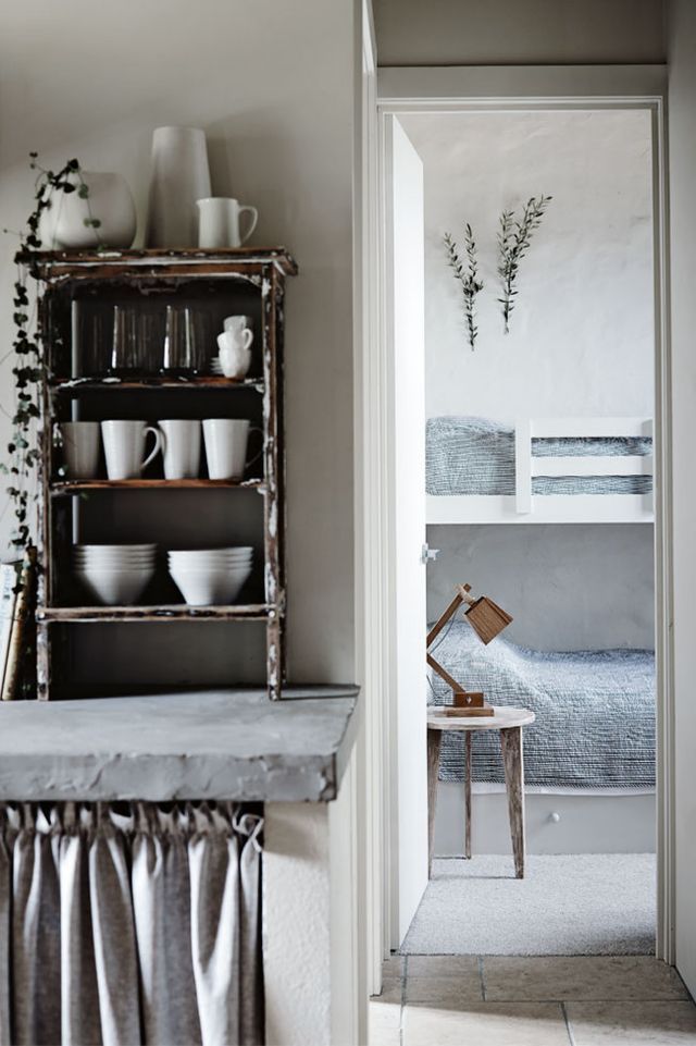 Charming kitchen with rustic shelving displaying white dishes, a concrete countertop, and a view into a cozy bedroom with soft blue bedding and minimalist decor.