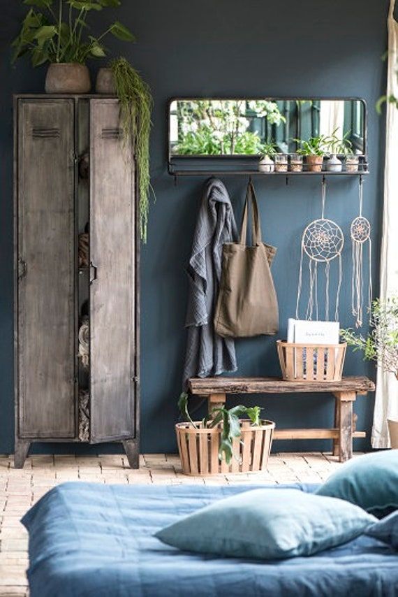A cozy bedroom featuring a dark blue accent wall with a vintage metal locker, a wooden bench with plants, and a mirror shelf with hanging dreamcatchers. The bed is dressed in blue linens and pillows, creating a calm and inviting atmosphere.