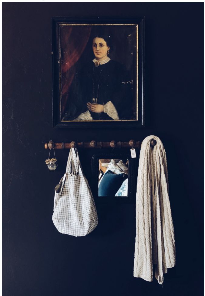 A dark, cozy entryway with a black wall, adorned with a classic portrait painting. Below the painting is a wooden hook rack holding a white checkered bag and a beige knitted scarf.