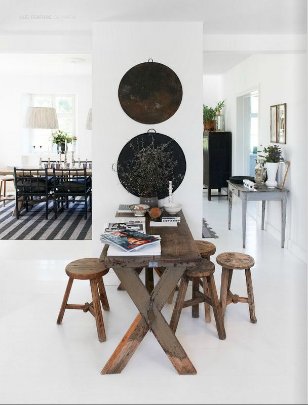 A cozy dining nook featuring a long wooden table with matching rustic stools. The walls are decorated with round, black metal wall hangings and a vase with dried branches as a centerpiece. The space exudes a simple, bohemian charm with a mix of natural materials and minimalistic decor, set against a bright, white background.