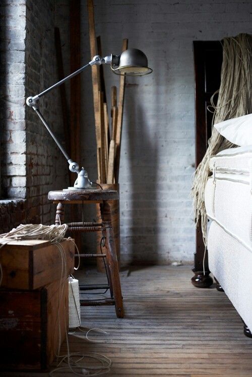 Rustic bedroom corner with a vintage adjustable metal desk lamp on a wooden stool, against a backdrop of exposed brick walls and scattered wooden planks. The setting exudes an industrial and minimalist vibe with earthy tones and textures.