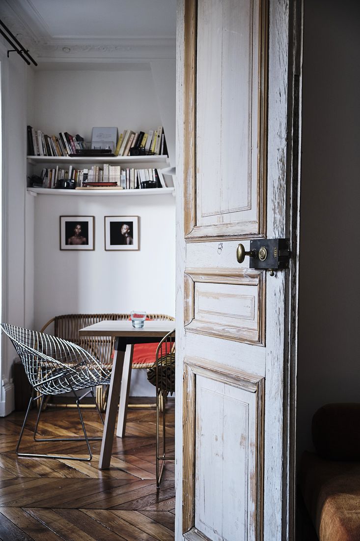 A cozy Parisian room with a rustic wooden door, shelves filled with books, a small table with chairs, and framed art on the walls.