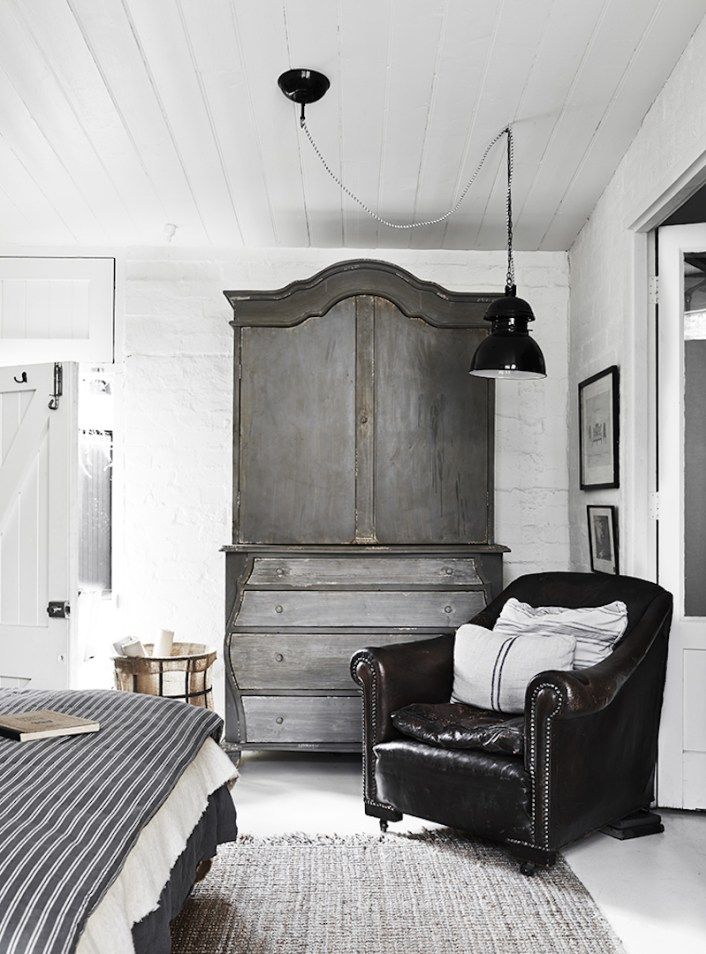 A stylish room with a dark leather armchair, a large rustic wardrobe, a cozy bed with striped linens, and minimalist decor.