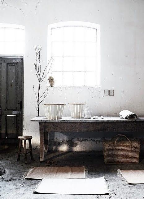 Rustic interior with a weathered wooden table, wicker baskets, white ceramic containers, and a small wooden stool, set against a backdrop of a whitewashed wall and large windows.