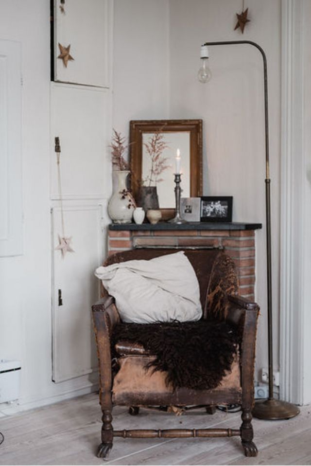 A charming corner of a home with a vintage armchair, a fluffy cushion, a floor lamp, and a small mantle with decorative items and a candle.