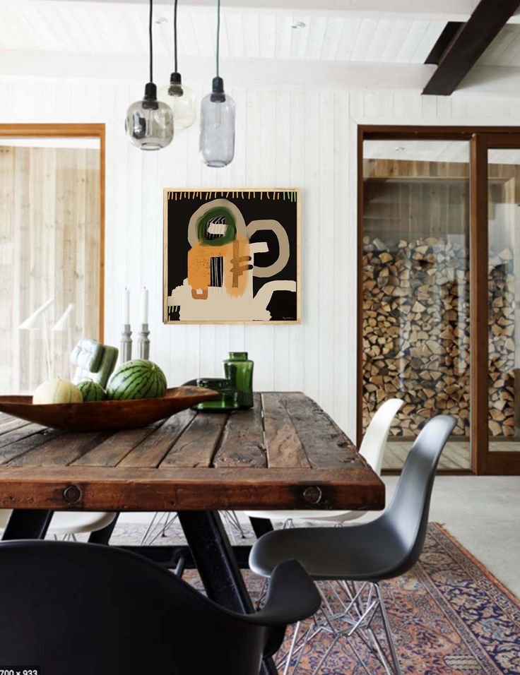 A dining area with a rustic wooden table and modern black and white chairs. The space is adorned with a framed minimalistic mid-century art print in natural colors on the wall. Overhead, pendant lights with glass shades provide illumination, and a bowl of watermelons sits on the table, adding a touch of freshness. A large pile of neatly stacked firewood is visible through the sliding glass doors in the background.