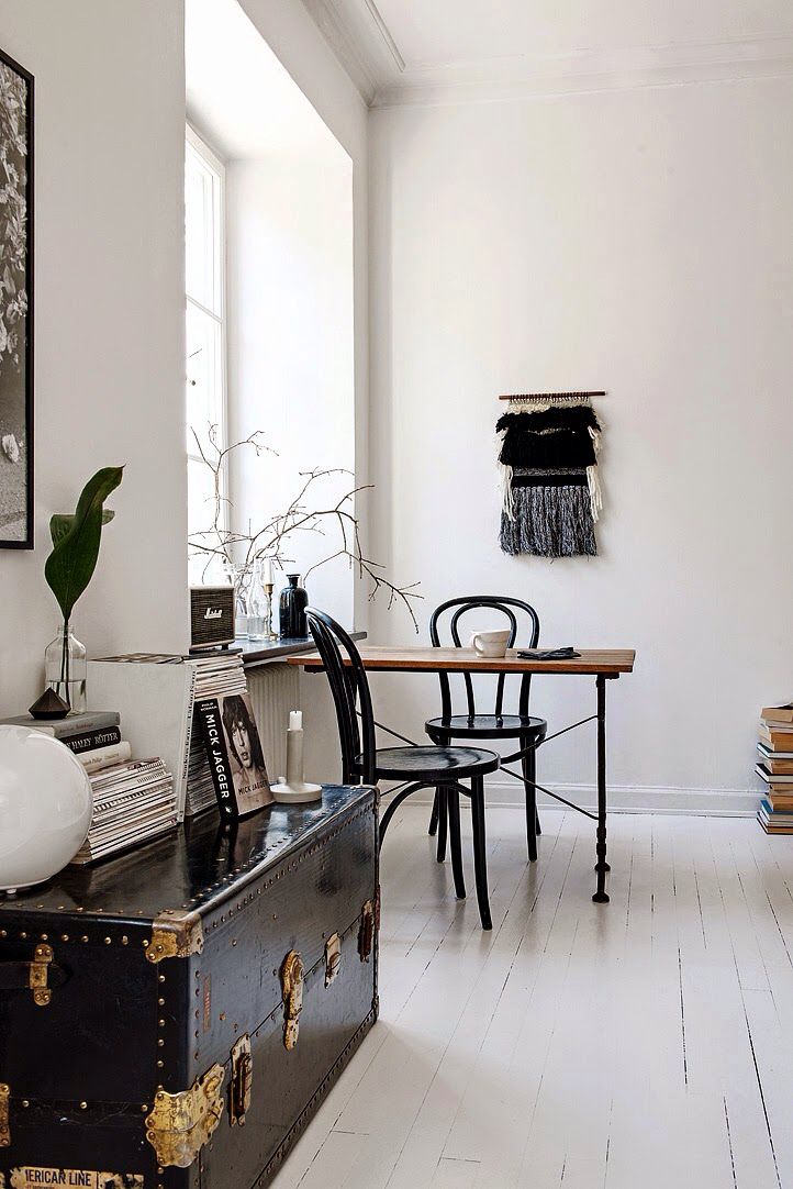 A bright and airy room with a minimalist aesthetic. The space includes a wooden table with black chairs, a vintage trunk, and a wall hanging in neutral tones. A mix of modern and vintage elements create a balanced and serene environment.
