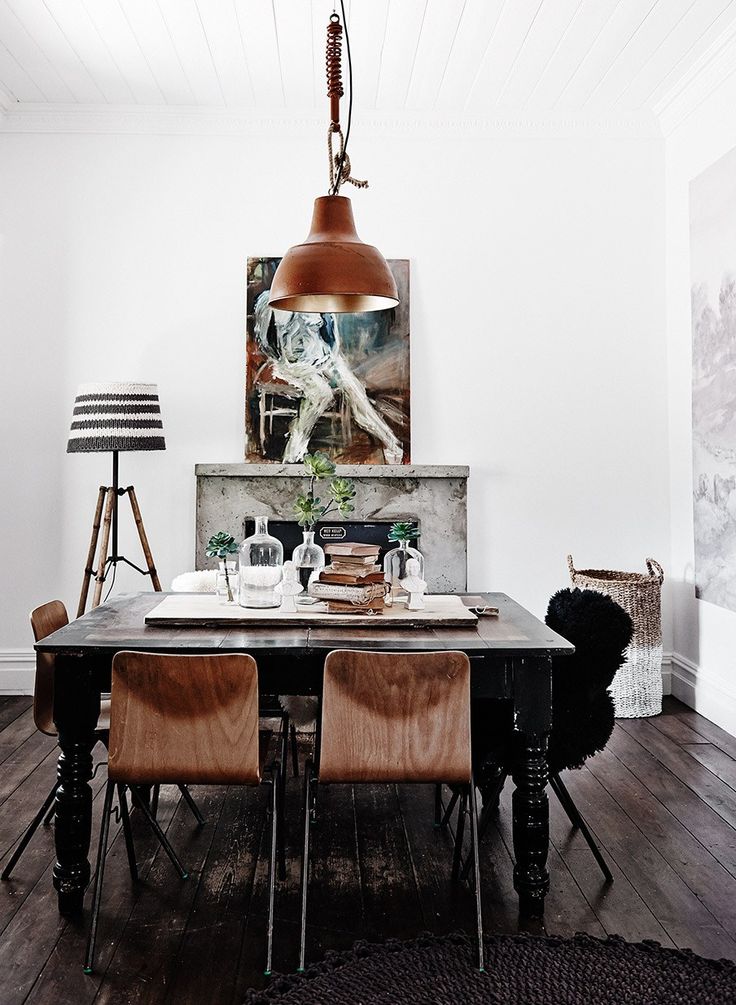 Rustic dining room with a large industrial-style pendant light hanging above a dark wooden table set with books and glass bottles, accompanied by wooden chairs. A floor lamp with a striped shade and a large painting add character to the space.