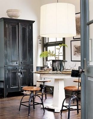 Country-style dining area featuring a round white table with wooden stools, a large white pendant lamp, and a tall black cabinet in the background, complemented by rustic decor and natural elements.
