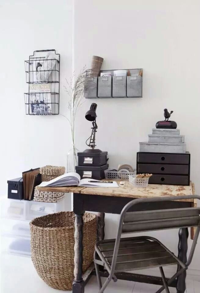 A cozy and organized home office corner with a wooden desk, a metal chair, and various storage solutions. The desk is adorned with a black lamp, small storage boxes, and a few decorative items. Wall-mounted wire baskets provide additional storage.