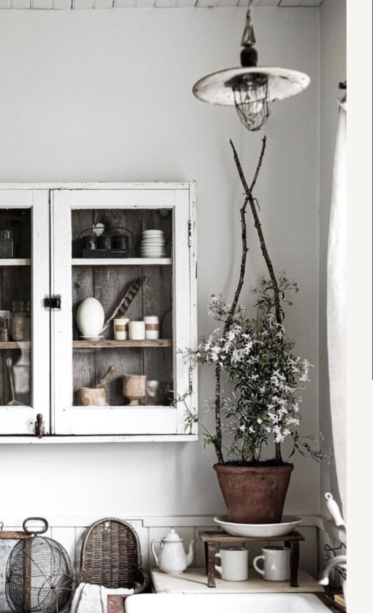Vintage kitchen with a rustic pendant light featuring a metal shade and exposed bulb, hanging above a countertop. The scene includes a white cabinet with glass doors displaying dishes, a potted plant, and various vintage kitchen accessories.