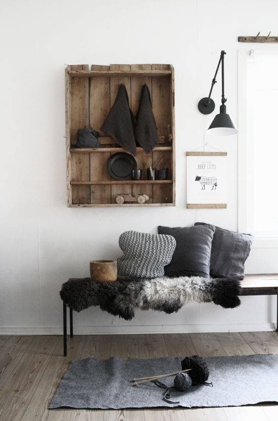A minimalist living space with a rustic wooden shelf, dark gray pillows, and a bench covered with fur throws. The decor includes industrial-style lighting and natural wood accents, adding a cozy and modern touch.