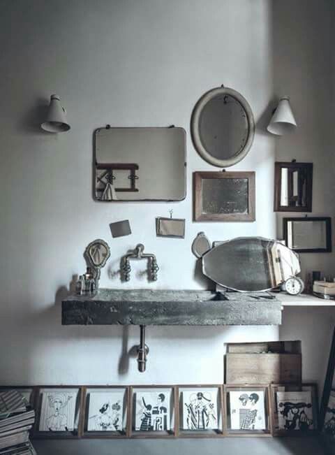 A vintage-style bathroom with an eclectic mix of mirrors in various shapes and frames. A concrete sink and faucet are paired with minimalistic wall lights and framed black-and-white artwork leaning against the wall.
