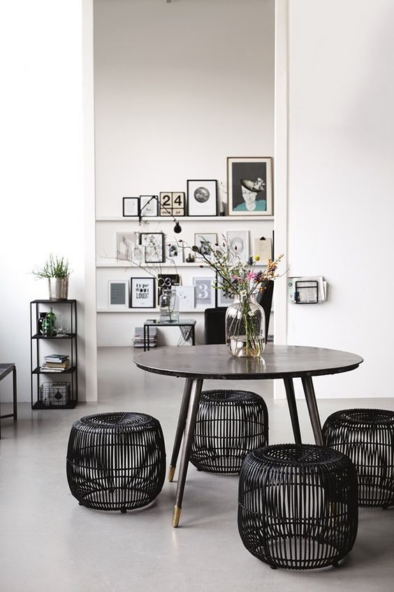 A contemporary dining area with a round wooden table surrounded by black rattan stools, featuring a minimalist decor with framed art on the wall and plants for a touch of greenery.