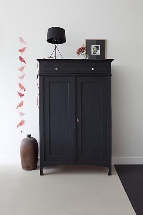 A sleek, modern black cabinet with two doors and a drawer at the top, adorned with a small black table lamp and a picture frame with flowers on top. A tall, round ceramic vase stands beside it on the floor, with a string of pink paper butterflies hanging on the wall to the left.