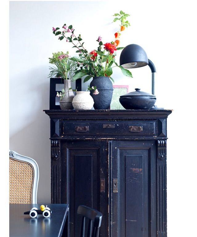 An antique black cabinet with two doors and intricate detailing, featuring some wear and tear. The top of the cabinet is decorated with a variety of vases, some with fresh flowers, and a black desk lamp with a curved neck.