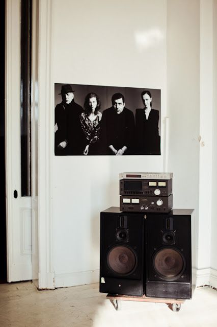 A minimalist room with a black and white poster of a rock band on the wall above a vintage stereo system and large speakers. The setup exudes a retro rock and roll vibe, with the stereo equipment placed on the floor, creating a simple yet striking focal point.