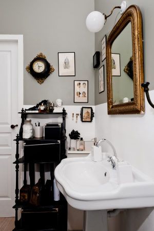 A classic bathroom featuring a white pedestal sink, an ornate gold-framed mirror, and black shelving. The wall is decorated with a collection of vintage-style framed prints and a black vintage clock.