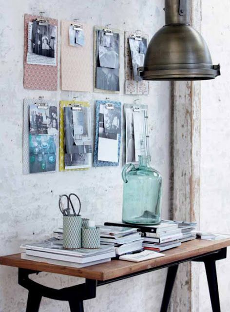 A minimalist home workspace with a rustic wooden desk covered in stacked books, small containers, and a glass bottle. The wall behind features a creative display of clipboards with black-and-white photos, under an industrial metal pendant light.
