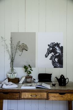 Rustic wooden desk with black and white botanical and horse artwork on the wall, a small potted plant, teapot, and open book, creating a serene and artistic workspace vignette.
