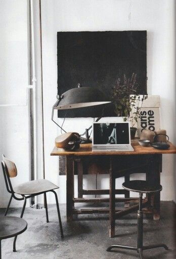 A rustic, industrial-style desk setup featuring a weathered wooden table with a black metal desk lamp and various decor items, including a vintage chair and stool. A laptop with a black and white screen is centered on the desk, with dark and moody art pieces displayed in the background, creating a rock and roll-inspired workspace.