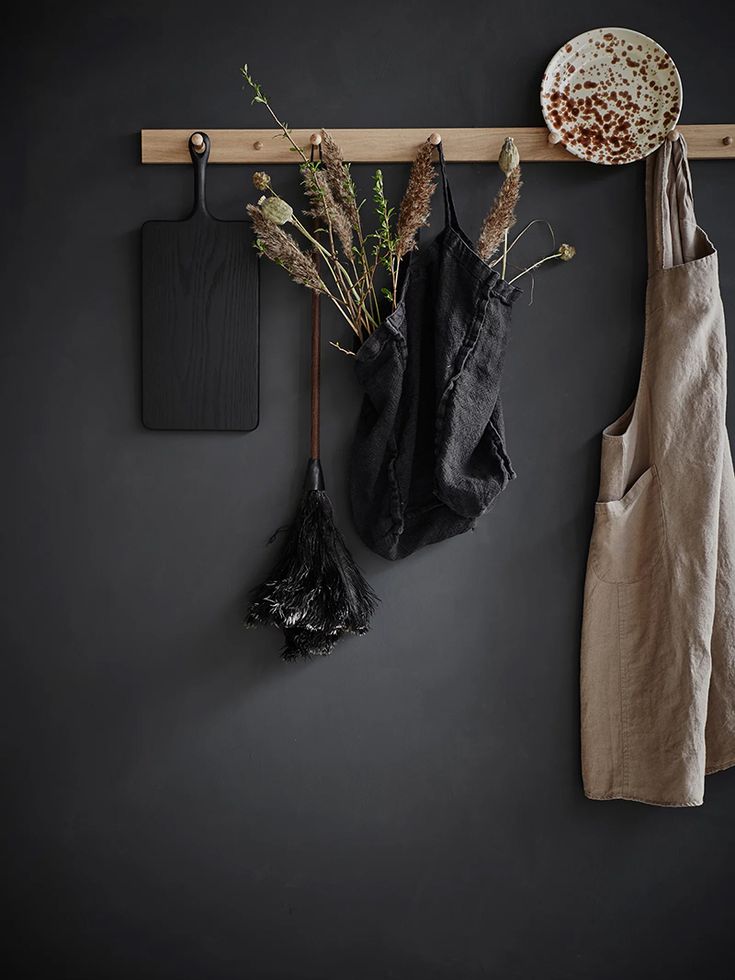 A simple, dark-themed interior featuring a matte black wall with a wooden hook rail. Aprons and natural decor items like dried grasses hang from the rail, accompanied by a cutting board and a speckled ceramic plate, creating a rustic and minimalist look.