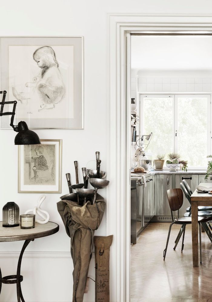 A cozy living area with a vintage feel, featuring framed sketches on a white wall, a small rustic table with decorative items, and a bundle of tools wrapped in canvas leaning against the wall. A view into the kitchen reveals natural light streaming through a large window.
