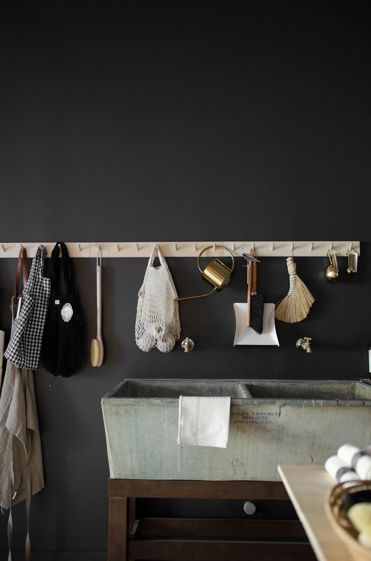 A modern utility space with a dark grey wall, featuring a row of hooks holding aprons, bags, and cleaning tools. A metal sink basin sits below, with brass accents and minimalist design elements contributing to a functional yet stylish setup.