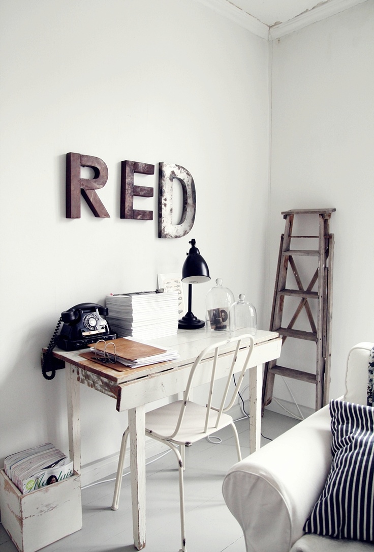 A minimalist white home office setup featuring a distressed wooden desk with a black retro telephone, a stack of magazines, a black desk lamp, and two glass cloche jars. The word ‘RED’ is displayed in large metal letters on the wall, with a rustic wooden ladder leaning against the wall