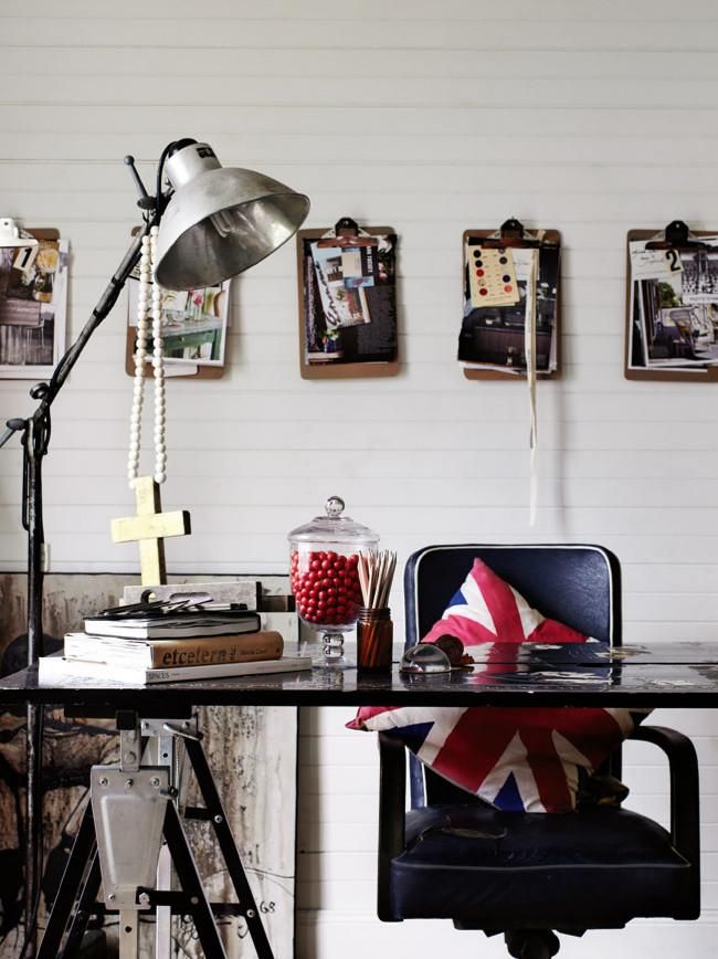 A stylish workspace featuring a vintage desk with a black industrial lamp, a jar of red candies, and a Union Jack pillow. The wall behind the desk is decorated with a row of clipboards holding various art and notes, creating an organized and inspiring display.