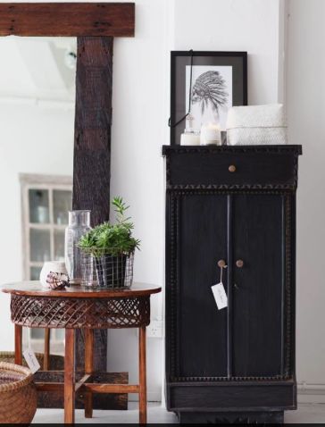 A narrow black cabinet with a carved design, standing next to a wooden round table with a wicker pattern. The cabinet is topped with candles, a small framed print, and a potted plant. A large rustic wooden mirror leans against the wall behind the table.
