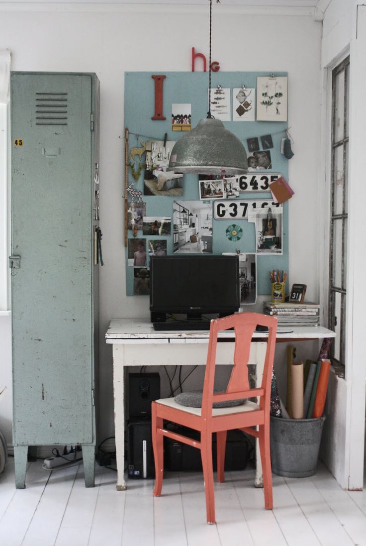 A vintage-inspired home workspace with a white distressed wooden desk, a coral-painted chair, and a green metal locker cabinet. The wall features an inspiration board with photographs, art, and ephemera, illuminated by a large industrial-style pendant lamp.