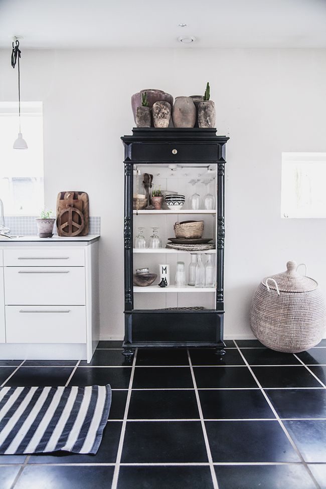 A modern kitchen with a black and white color scheme featuring a black display cabinet filled with rustic pottery, a striped rug on a black tiled floor, and minimalist white cabinets.